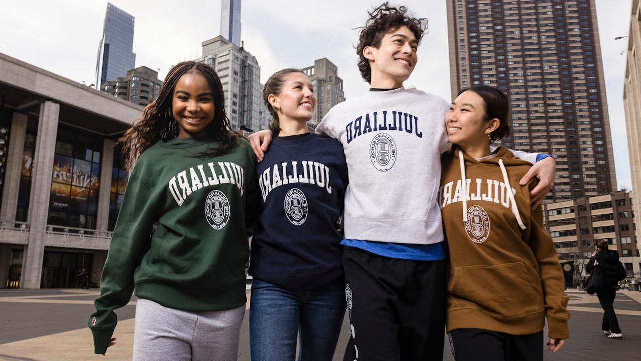 Four smiling students wearing Juilliard sweatshirts in different colors, standing arm-in-arm outdoors